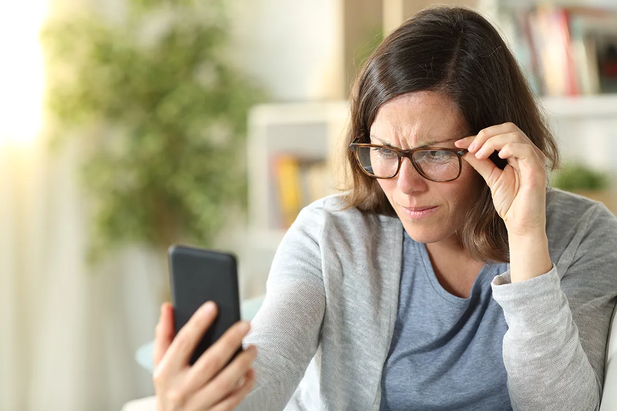 a woman sitting at home and having trouble seeing her phone with glasses on