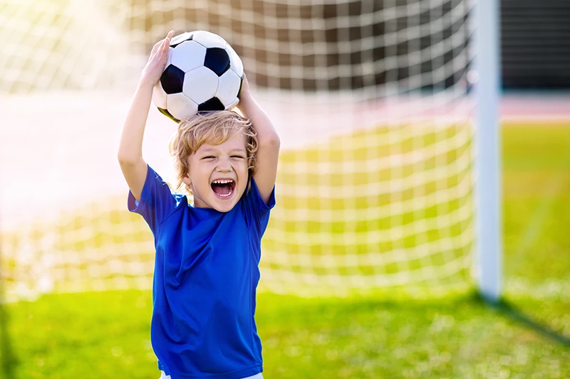 A little boy holding a soccer ball in front of a soccer goal outside.