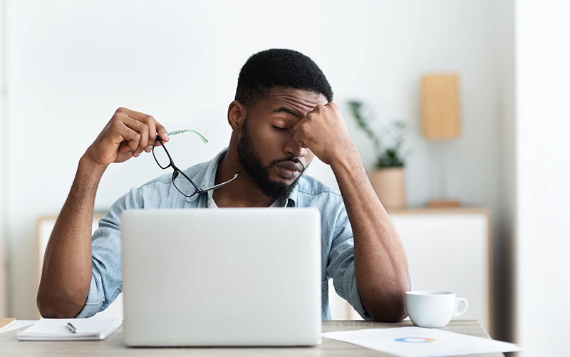 a man sitting at his computer taking off his glasses and resting his head on his hand